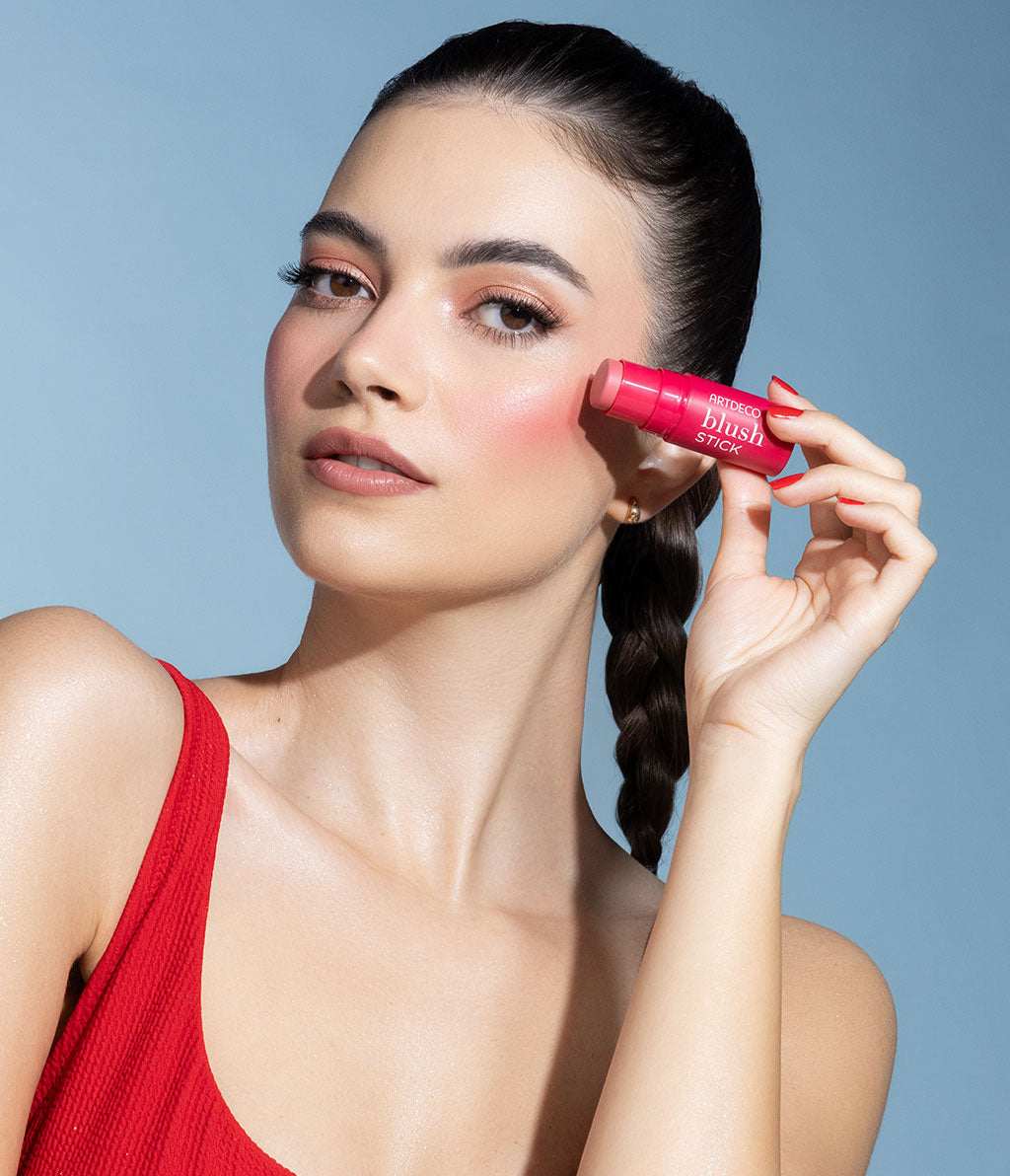 A woman with natural make-up holds a blush stick to her cheek and looks gently into the camera. She is wearing a red top and has braided hair.