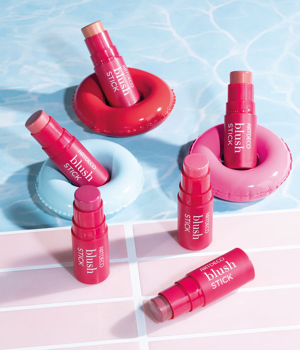 Six blush sticks in different colors are arranged on colorful floating rings that float on a water surface. The products exude freshness and summery radiance.