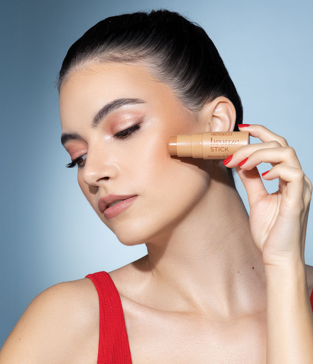 A woman with summery make-up holds a bronze stick to her cheek and looks to the side. She is wearing a red top and has tightly braided hair.