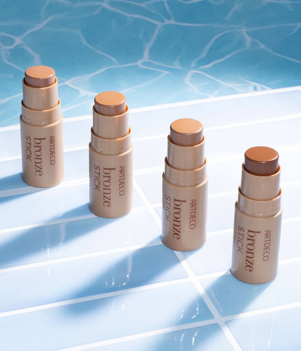 Four bronze sticks stand in a row on pool tiles while they are reflected on the surface of water. The products exude summery radiance.