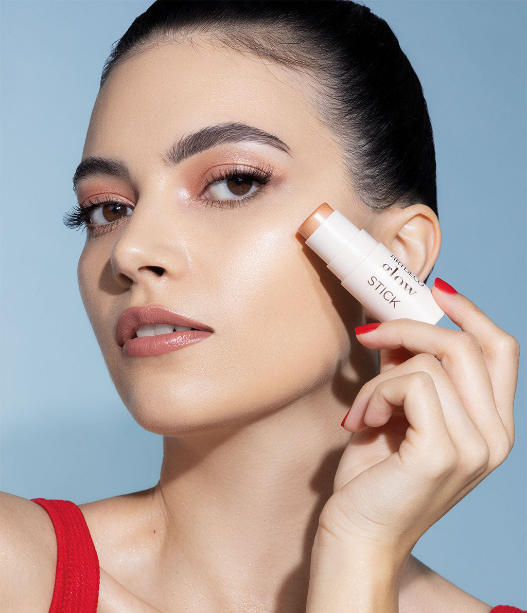 A woman with shimmering make-up applies highlighter to her cheekbones with an ARTDECO Glow Stick. Her look is summery-fresh with shining skin and subtle eyeshadow.