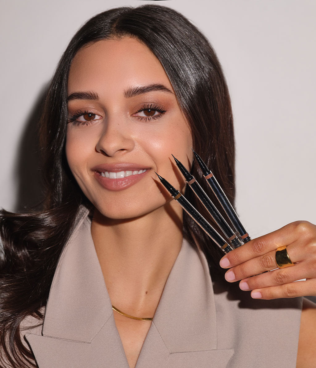 Brown-haired model holding the three new colors of the 24h Micro Brow Liquid Liner for perfect eyebrows