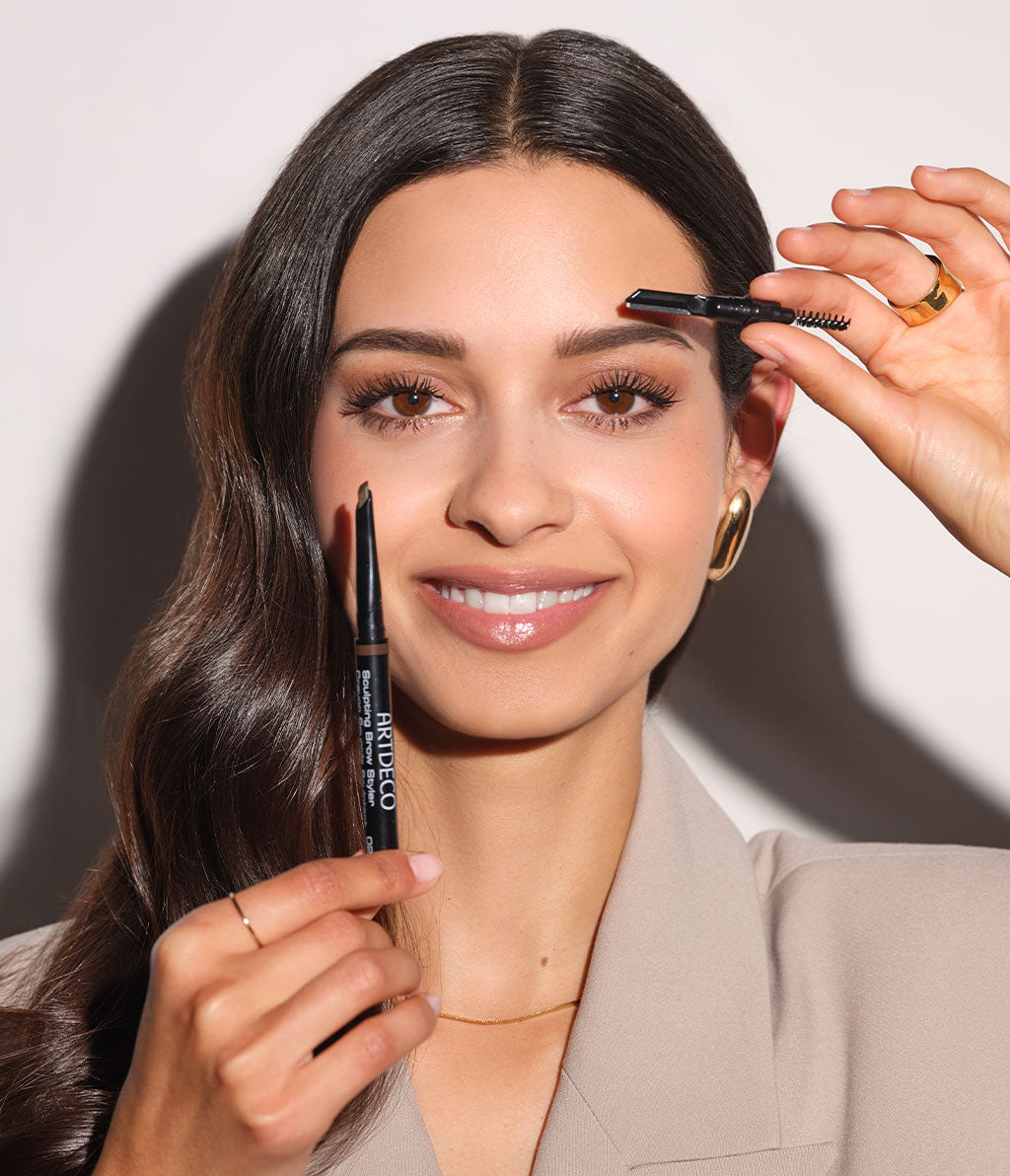 Brown-haired model holds the Sculpting Brow Styler with twisted-out refill in one hand. The 2-in-1 brush and eyebrow shaver are in the other hand