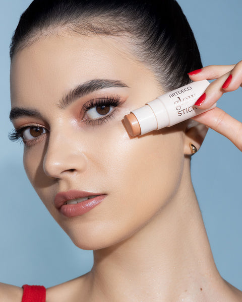 A woman with shimmering make-up applies highlighter to her cheekbones with an ARTDECO Glow Stick.