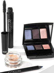 A palette, mascara, base, and kajal lie on a white background.