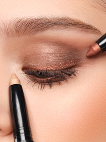 Two High Performance Eyeshadow Stylos in the colors gold and bronze are held to one eye
