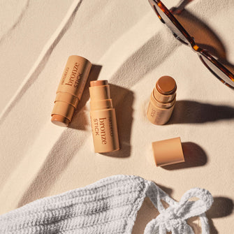 Bikini, sunglasses, and bronzer sticks in three colors placed on sand.