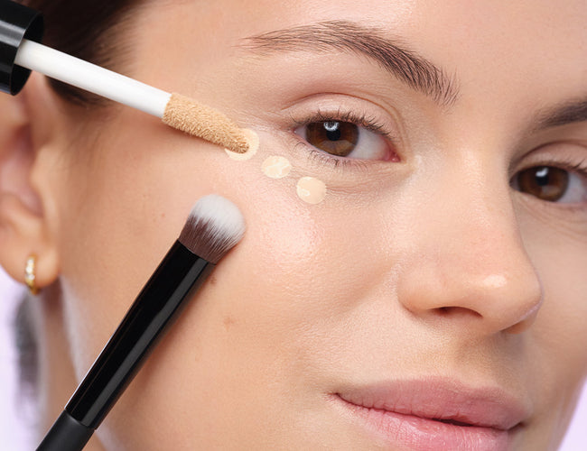 Close-up of the model’s eye, to which concealer has been precisely applied. The applicator and brush are also shown