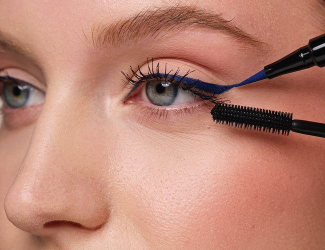Close up of the model’s eye, to which a blue eyeliner and black mascara are being applied.