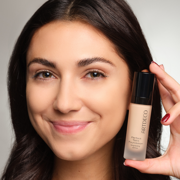 Model with a subtle makeup look holds a foundation next to her face