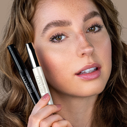 A model with mascara-coated eyelashes holds two different mascaras next to her face.