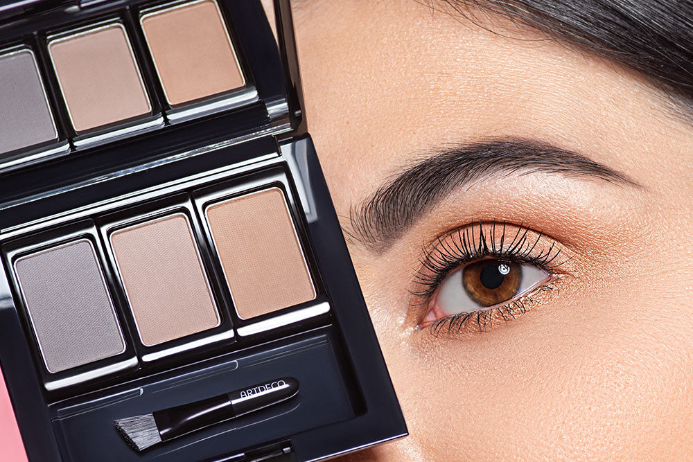 A woman with made up eyebrows is shown holding an eyebrow powder palette in front of her face