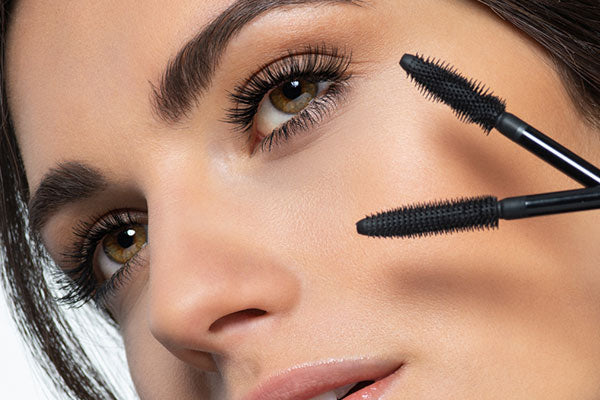 A woman with made up eyes holding two mascara brushes in front of her face