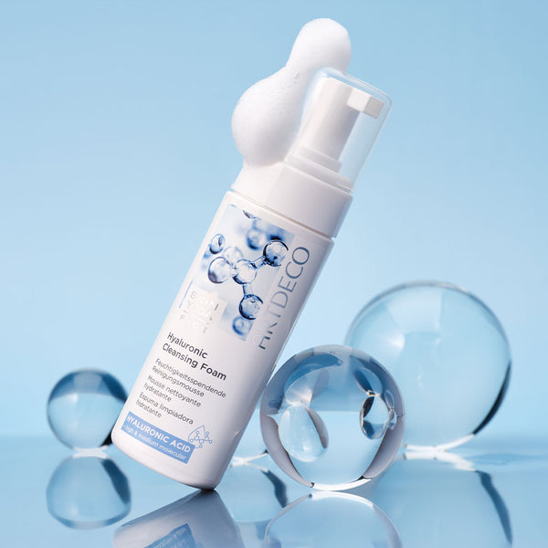 Hyaluronic Cleansing Foam on a light blue background surrounded by different-sized glass beads. Some foam can be seen on the lid.
