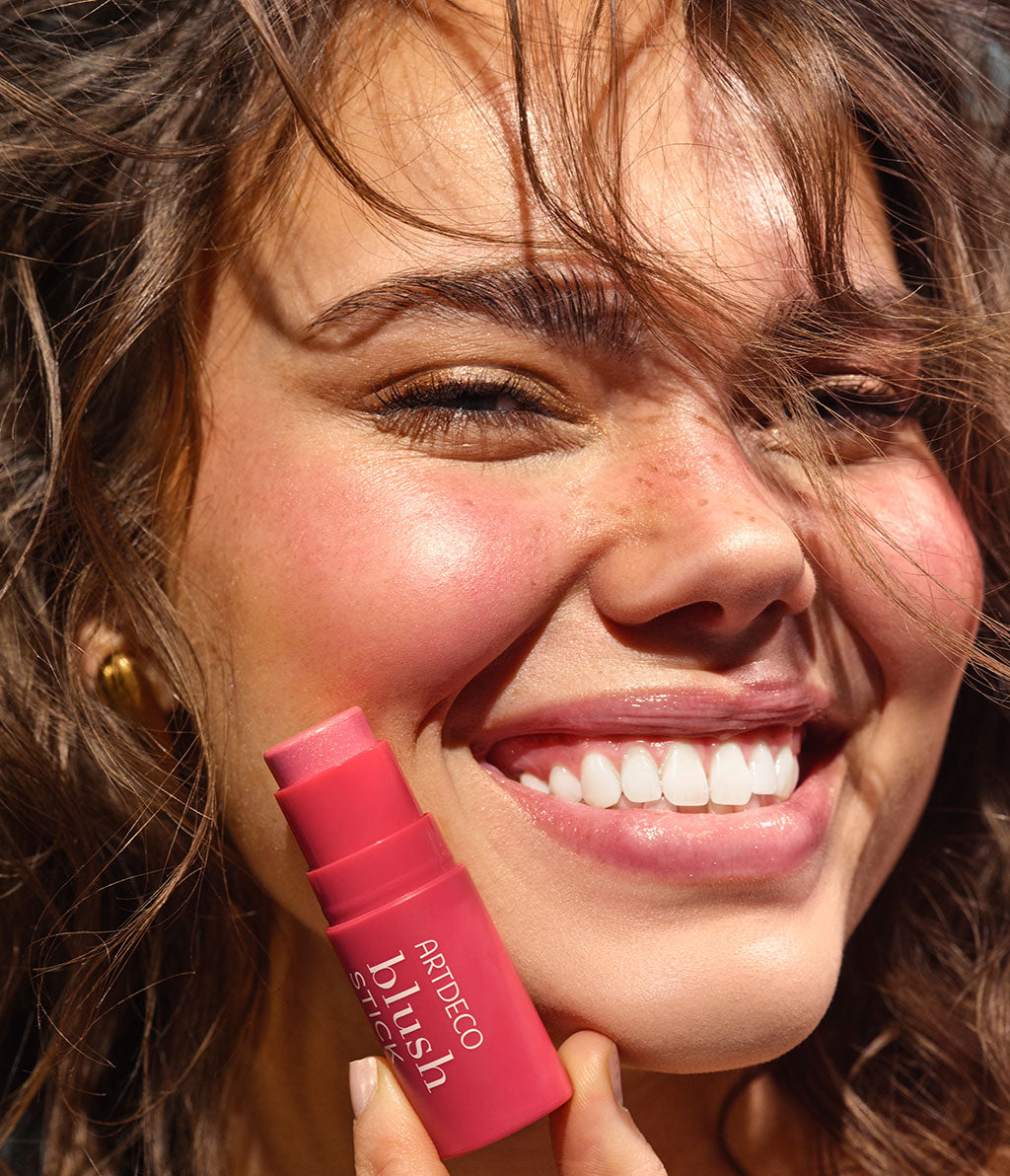 A young woman with curly hair smiles radiantly into the camera as she holds a blush stick in her hand and applies it to her cheeks.