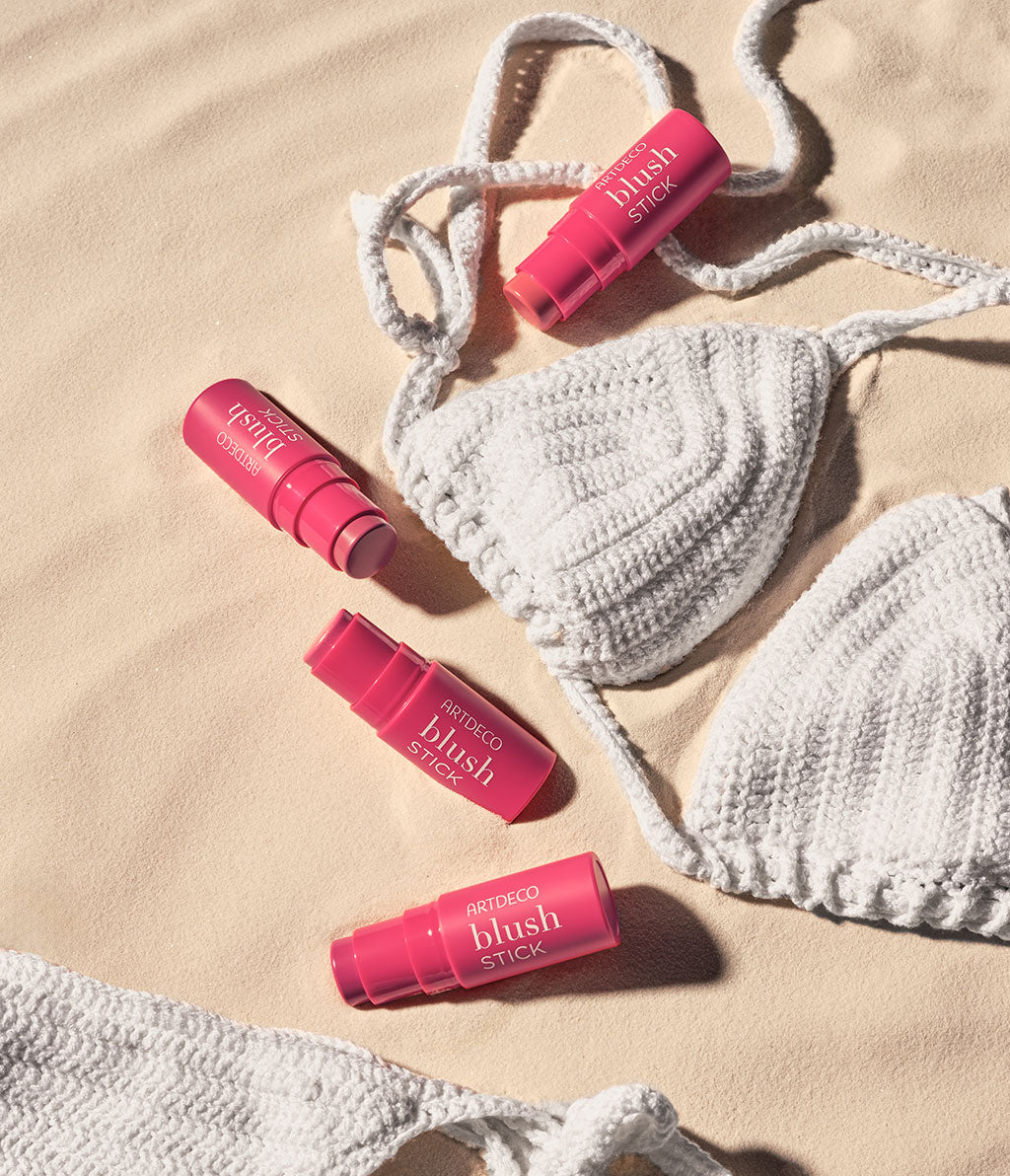 Several blush sticks from ARTDECO lie on a sand-colored background, surrounded by a white, knitted bikini.