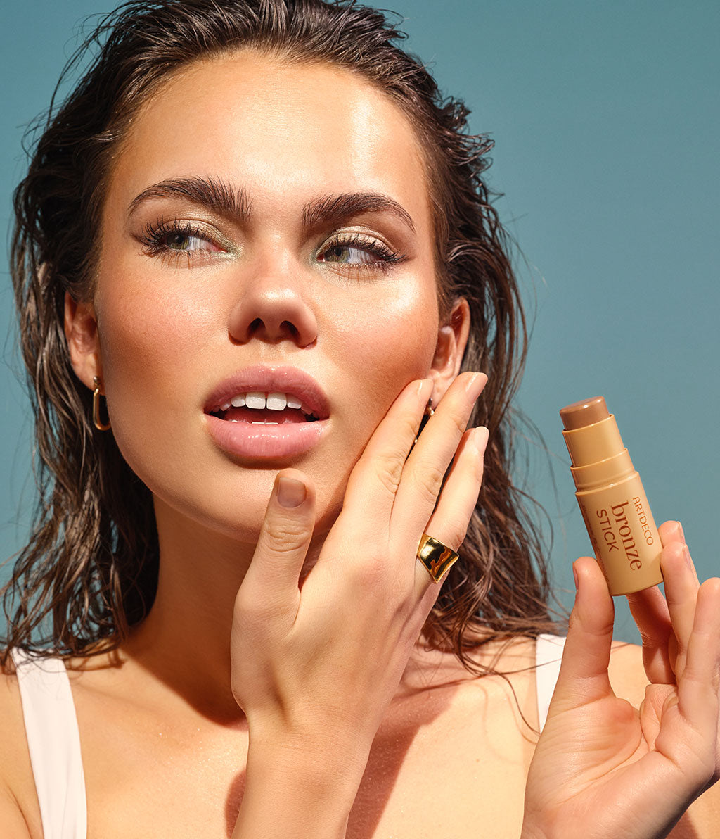 A young woman with curly hair smiles radiantly into the camera as she holds a bronze stick in her hand and applies it to her cheek with her fingers. She has a naturally tanned, summery complexion.