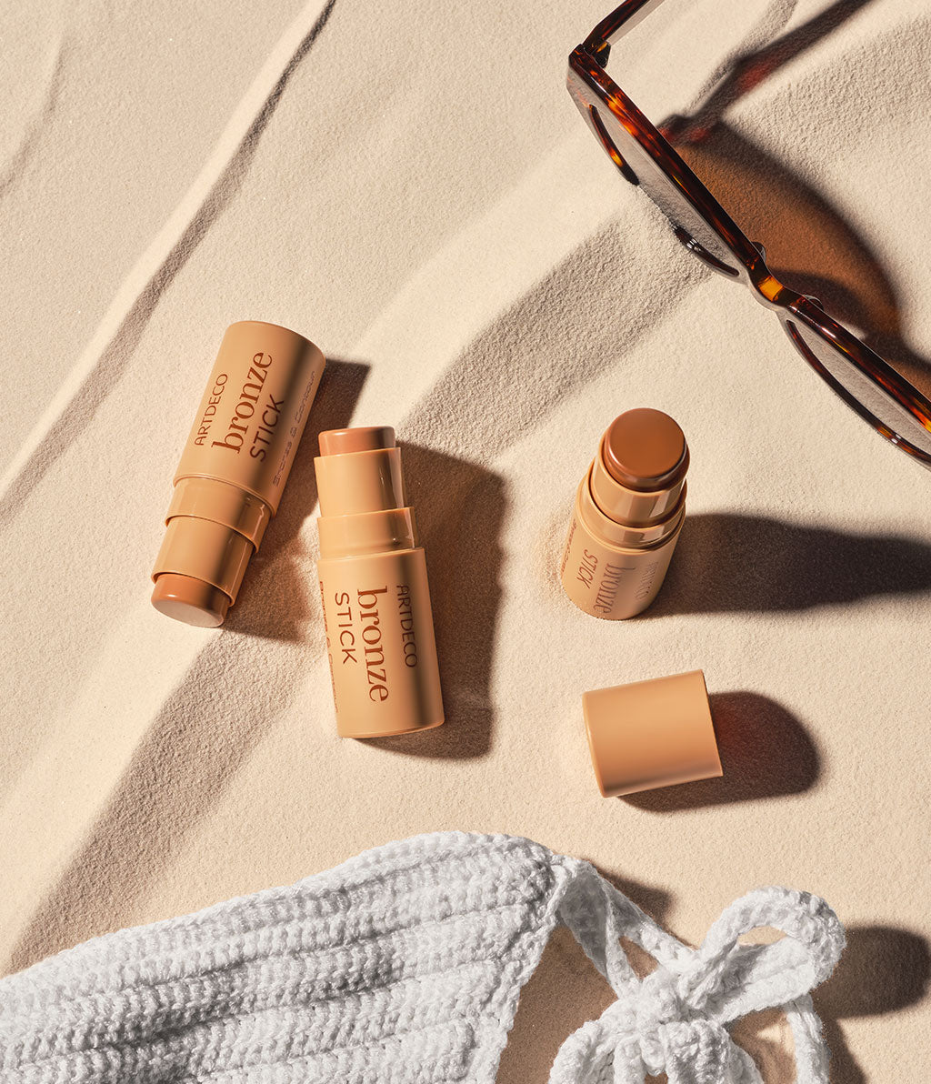 Several bronze sticks from ARTDECO lie on a sand-colored background, surrounded by a white, knitted bikini.