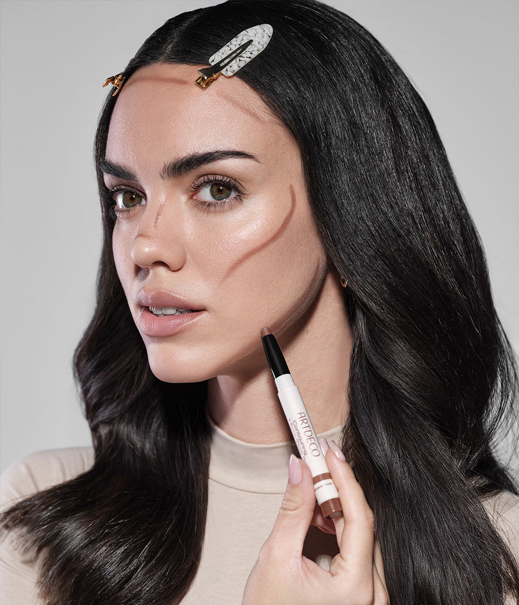 Brunette model holding the Contouring Micro Stick. The stick has already been applied to her forehead, nose, and cheeks, but has not yet been blended.