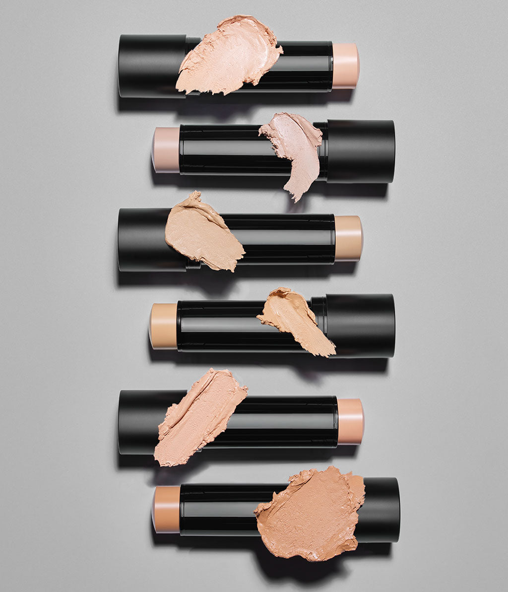 All six Skin Perfecting Face Sticks are lined up on a gray background. Each texture swatch is positioned over its corresponding stick.