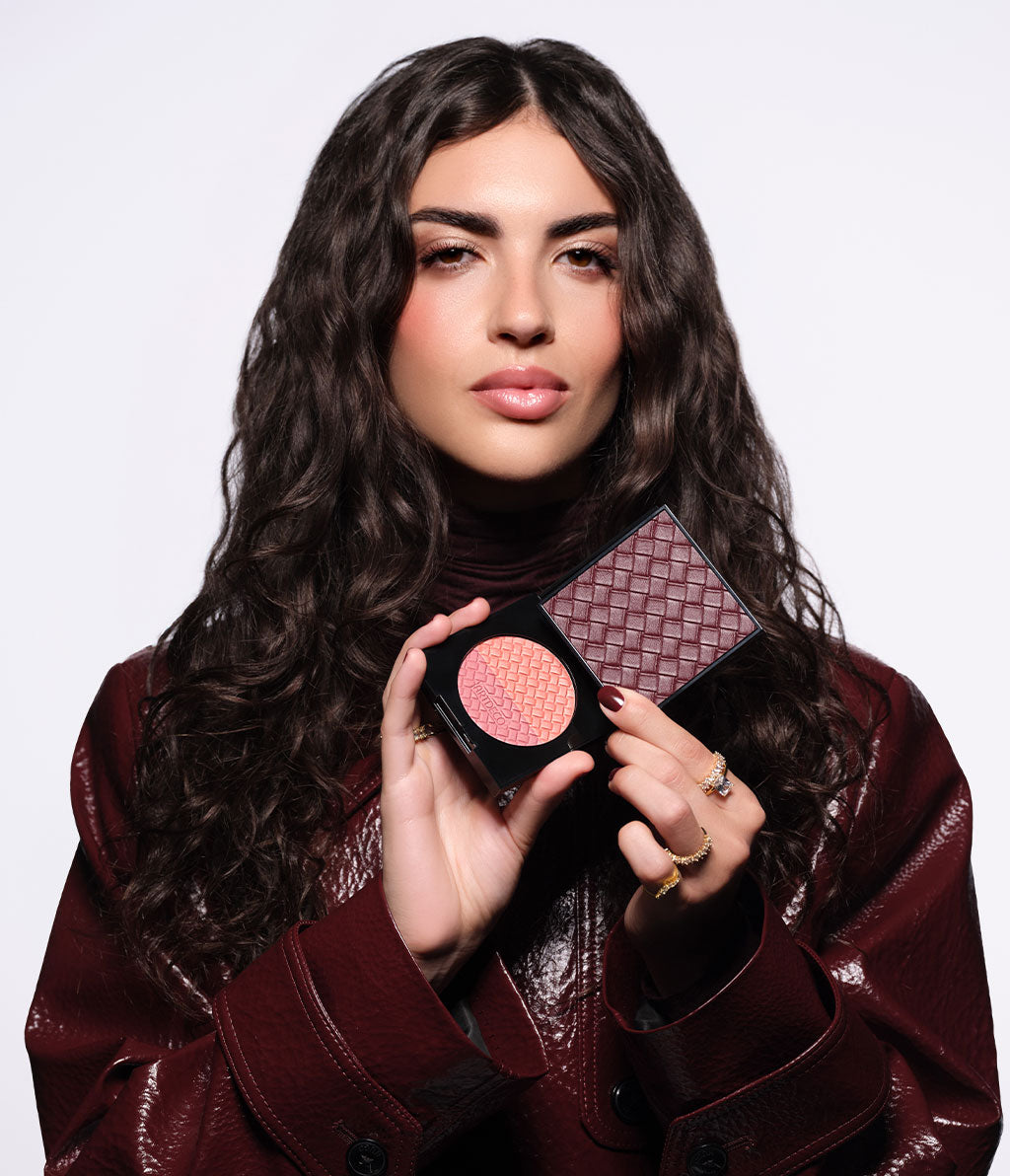A brunette model is wearing a burgundy jacket and holding the open Blush Couture in her hand, so that the embossing and the Intrecciato design are clearly visible.