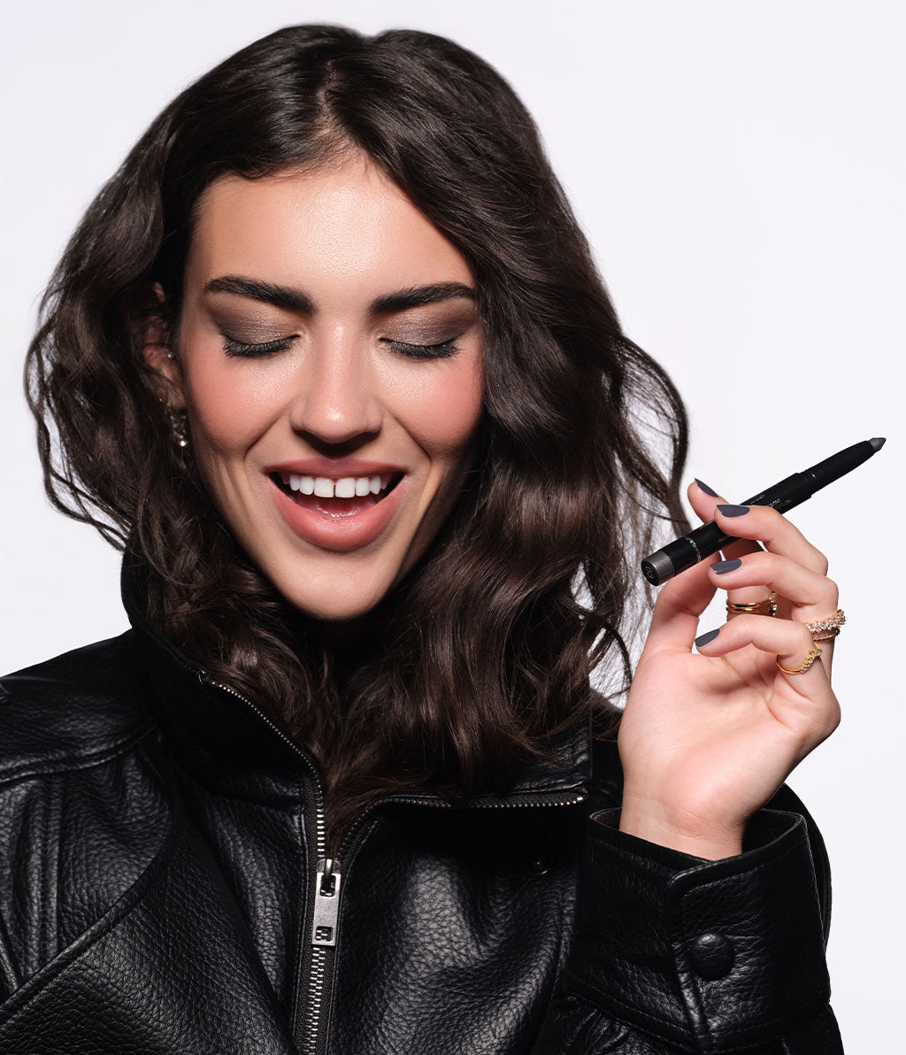 A brunette model wearing a black jacket and fall makeup is holding the High Performance Eyeshadow Stylo No. 02.