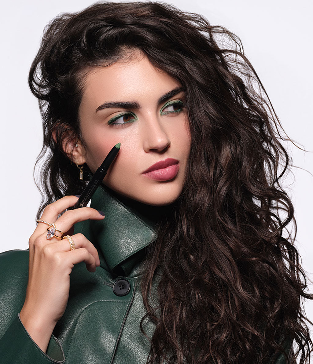A brunette model wearing a green jacket and fall-inspired makeup is holding the High Performance Eyeshadow Stylo No. 80.