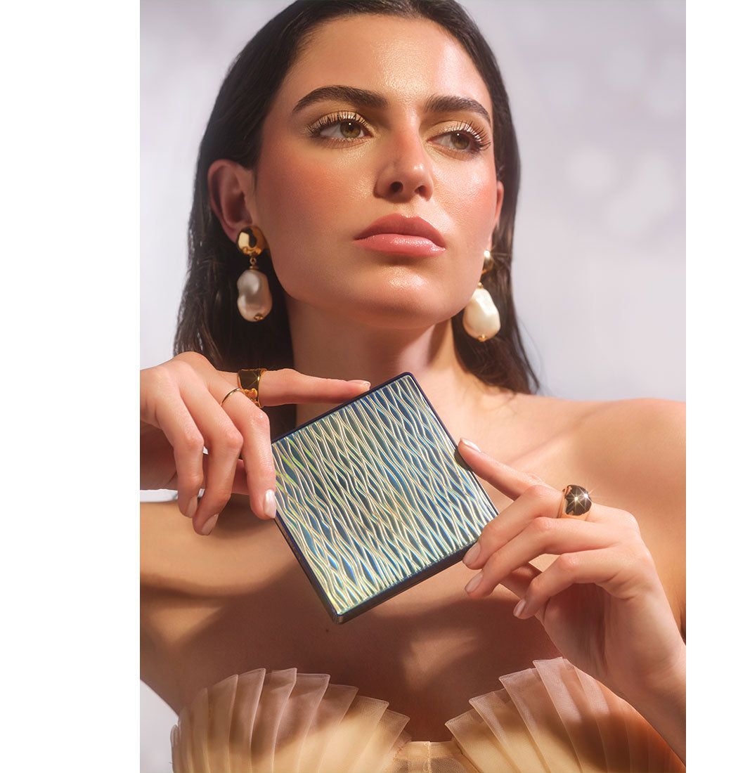 Brunette model wearing summery, glowing makeup and holding All Seasons Bronzing Powder in her hands to showcase its wavy packaging.