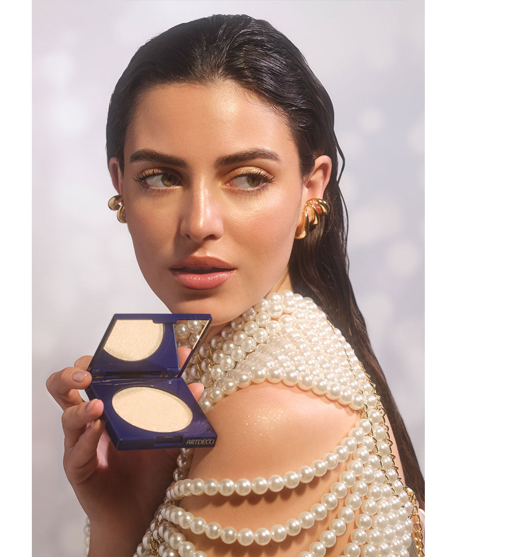 Brunette model looks over her shoulder while holding a partially opened Diamond Beauty Powder. The champagne-toned all-over highlighter is applied to both the face and shoulders to create an irresistible glow.