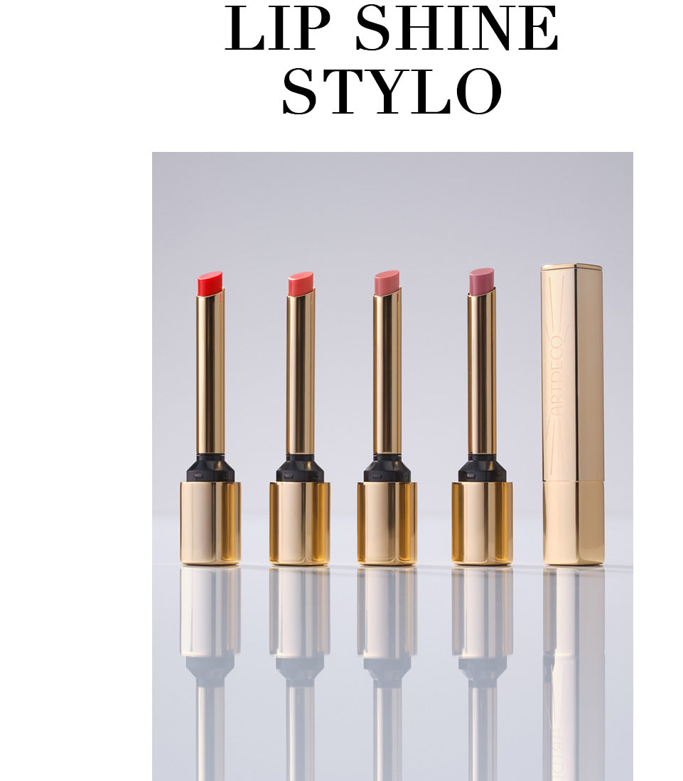 All four Lip Shine Stylo products are displayed open in a row against a neutral background. A fifth Lip Shine Stylo stands closed next to them. The delicate, gold packaging conveys the product’s high quality. The product name, “Lip Shine Stylo,” is also written in capital letters.