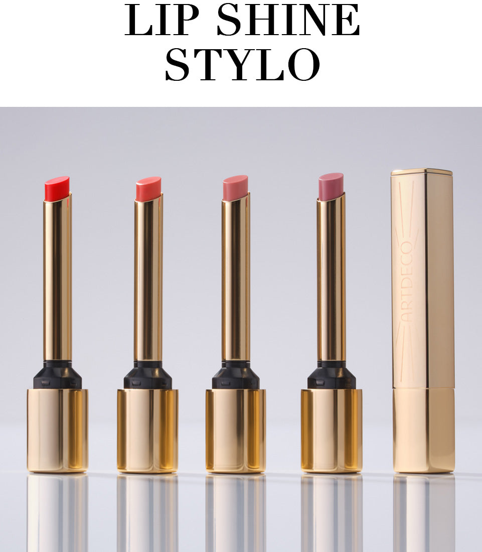 All four Lip Shine Stylo products are displayed open in a row against a neutral background. A fifth Lip Shine Stylo stands closed next to them. The delicate, gold packaging conveys the product’s high quality. The product name, “Lip Shine Stylo,” is also written in capital letters.