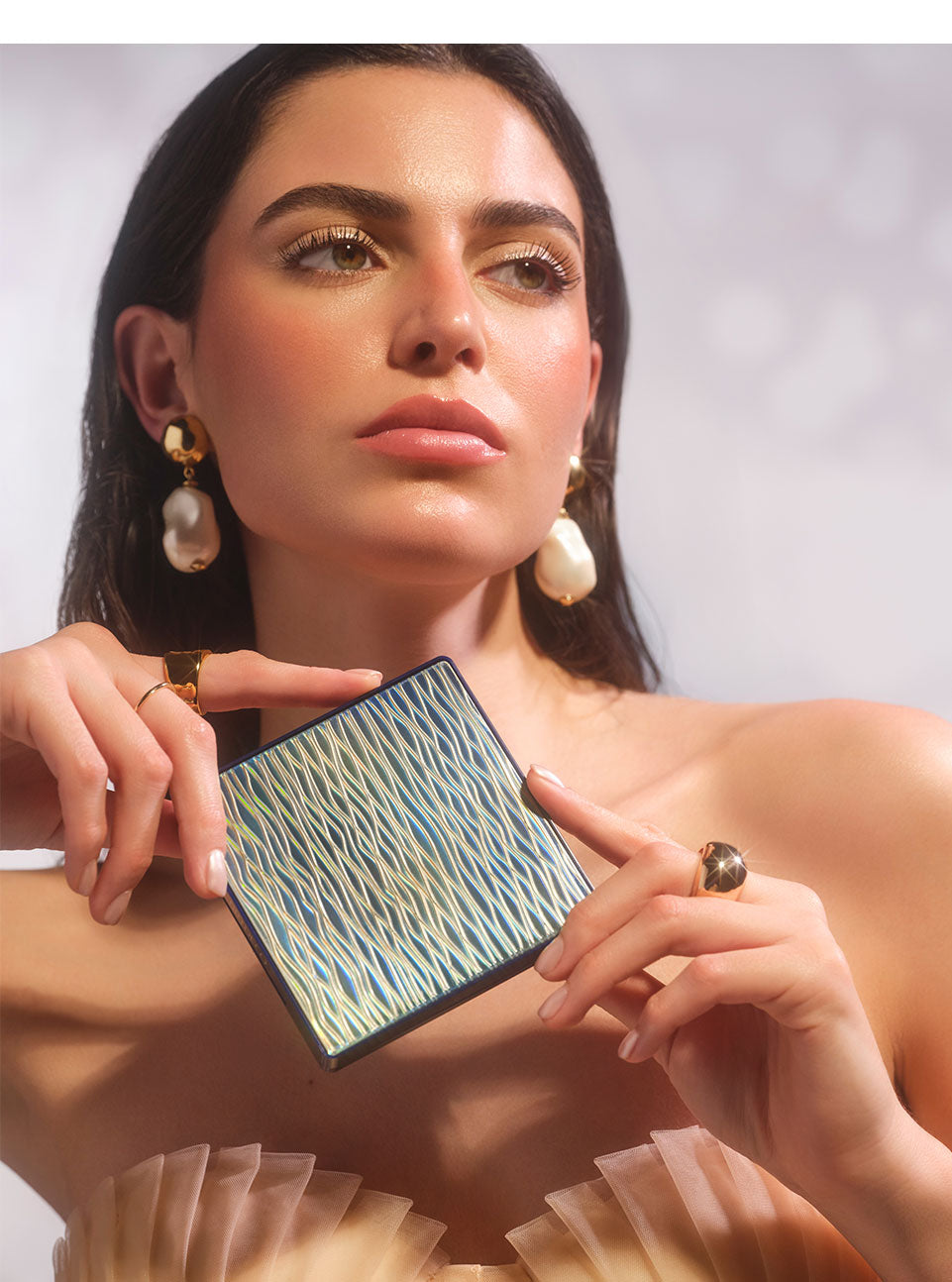 Brunette model wearing summery, glowing makeup and holding All Seasons Bronzing Powder in her hands to showcase its wavy packaging.