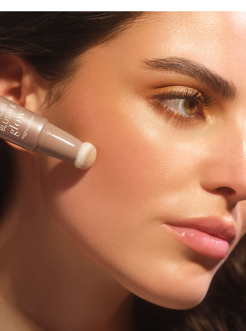 Brunette model applies the Wonder Blush & Glow Highlighter to her cheek with the sponge applicator to create a glowing makeup look.