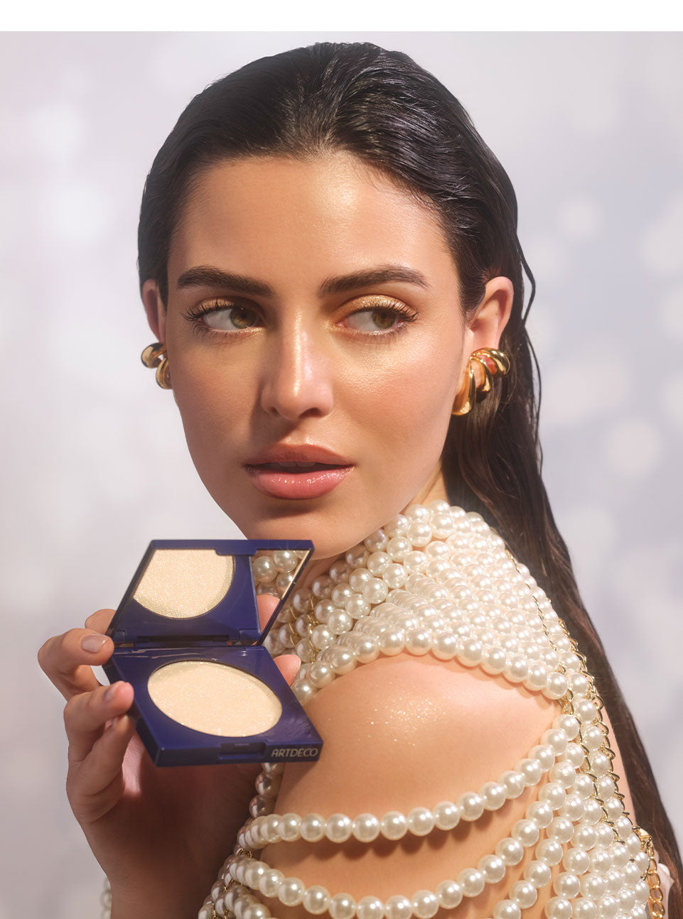 Brunette model looks over her shoulder while holding a partially opened Diamond Beauty Powder. The champagne-toned all-over highlighter is applied to both the face and shoulders to create an irresistible glow.