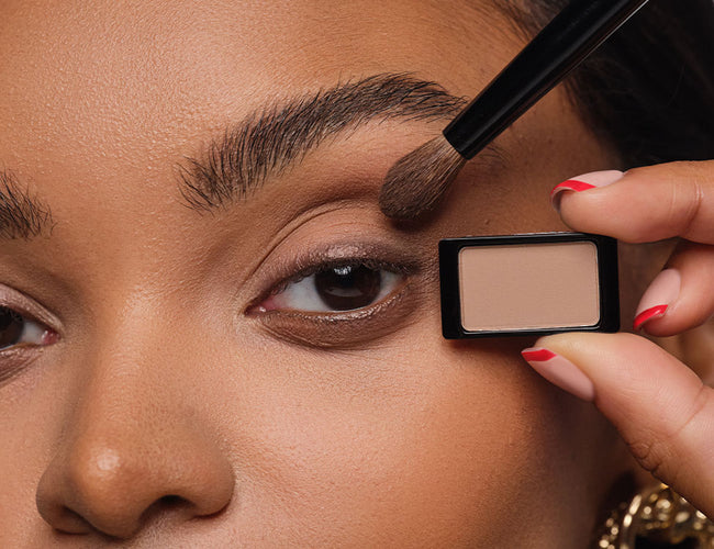 The model holds the neutral eyeshadow in her hand and shows how to apply it to the eye with a fluffy eyeshadow brush