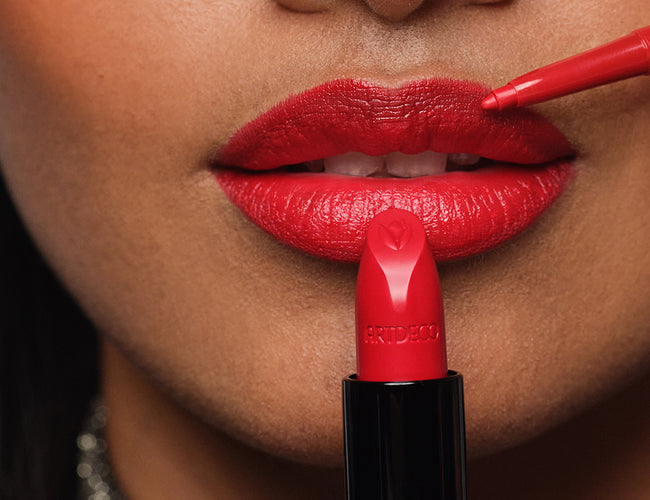 Close up on the model’s made-up lips, with a red lip liner and red lipstick held up to them 