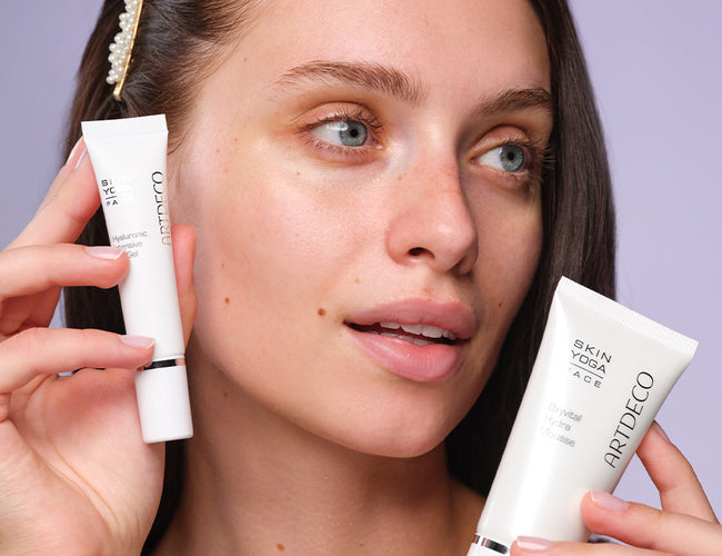 A dark-haired model holds two different ARTDECO skincare products in her hands and smiles while looking to the side.