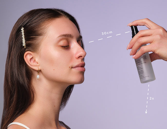 A dark-haired woman sprays 3-in-1 Makeup Fixing Spray on her face. Her eyes are closed and she has a calm expression on her face.