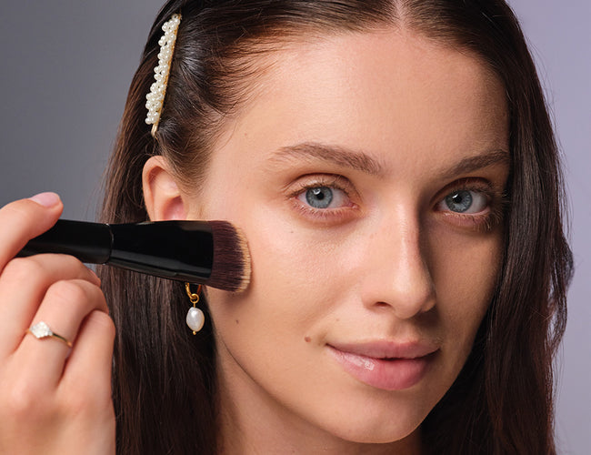 A dark-haired model uses a makeup brush to apply the bridal makeup foundation evenly across her face.