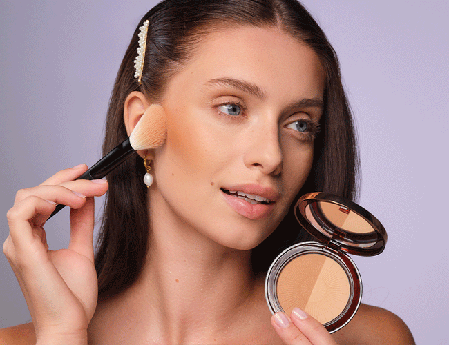 A dark-haired model holds Bronzing Powder in one hand while applying the powder to her cheek with a powder brush in the other.