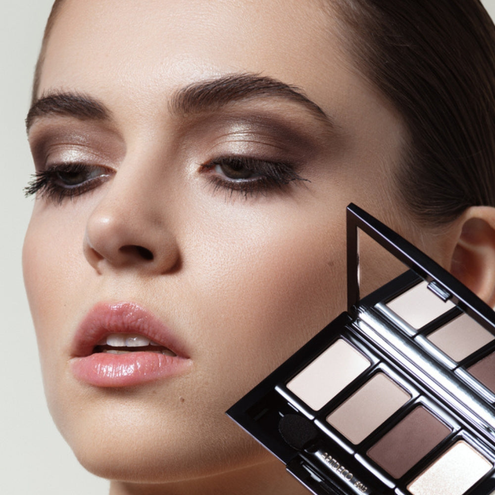 A woman with makeup on her eyes is holding a beauty box with eyeshadow in her hand.