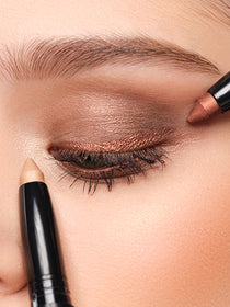 Two High Performance Eyeshadow Stylos in the colors gold and bronze are held to one eye 