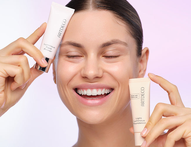 A smiling model holds the foundation and primer for everyday makeup with a natural glow in each hand 