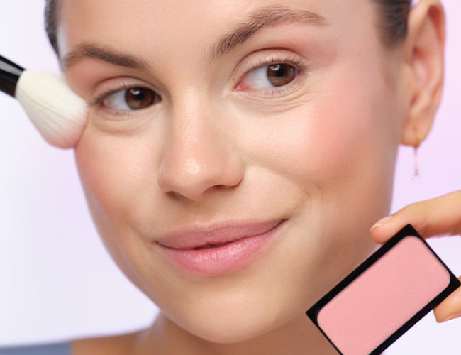 Model applies the pink blush using a powder brush, which she holds in her other hand