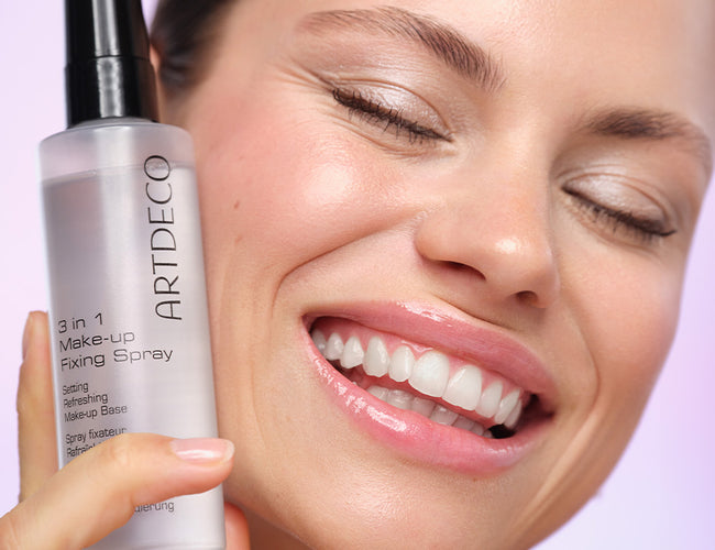 The model is smiling broadly with her eyes closed and holds the Fixing Spray to her cheek.