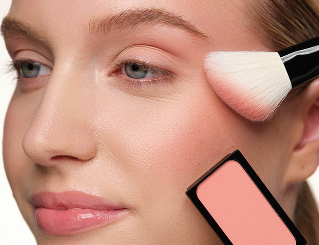 Close-up of the model’s cheeks, to which a pink blush is being applied using a blush brush