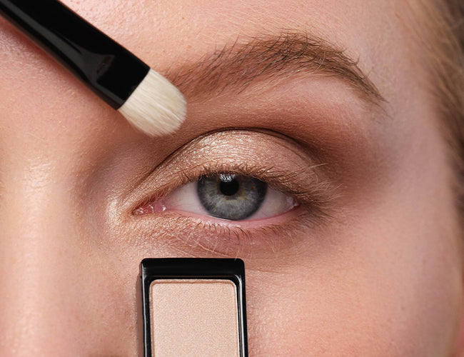 Close-up of the model’s eye, to which a Champagne-colored eyeshadow is being applied to the inner eye corner 