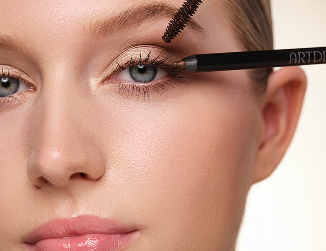 Close-up of the model’s eye with the finished makeup look, to which a brown mascara and a brown eyeliner are held up at the same time