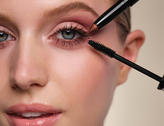 Close-up of the model’s eye with the finished makeup look, to which a black mascara and a copper-colored eyeshadow are held up at the same time