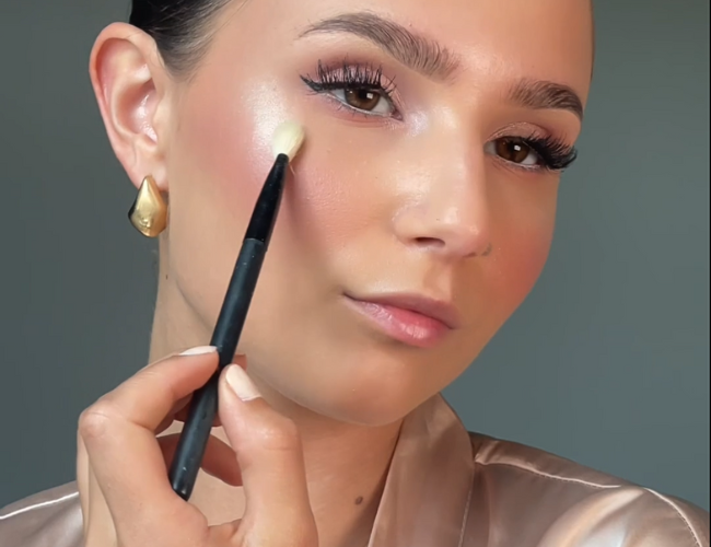 The highlighter is applied to the upper cheekbones with a small brush.